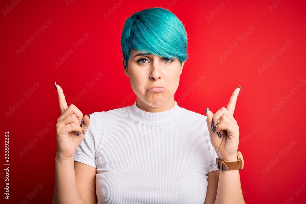 Young beautiful woman with blue fashion hair wearing casual t-shirt over red background Pointing up looking sad and upset, indicating direction with fingers, unhappy and depressed.