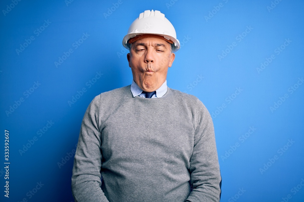 Middle age handsome grey-haired engineer man wearing safety helmet over blue background making fish face with lips, crazy and comical gesture. Funny expression.