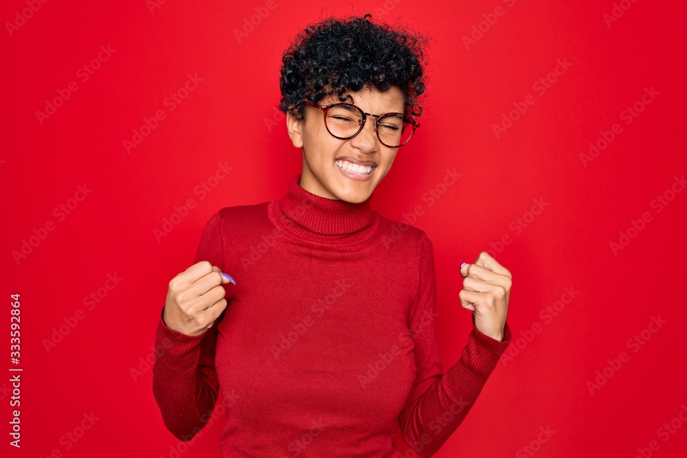 Young beautiful african american afro woman wearing turtleneck sweater and glasses very happy and excited doing winner gesture with arms raised, smiling and screaming for success. Celebration concept. Young beautiful african american afro woman wearing turtleneck sweater and glasses very happy and excited doing winner gesture with arms raised, smiling and screaming for success. Celebration concept.