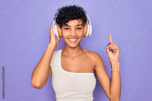 Beautiful african american woman listening to music using headphones over red background surprised with an idea or question pointing finger with happy face, number one
