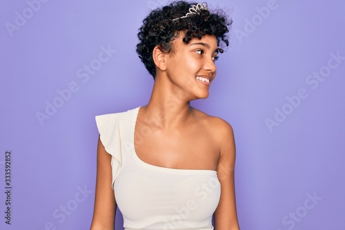 Young beautiful african american afro woman wearing tiara crown over purple background looking away to side with smile on face, natural expression. Laughing confident.