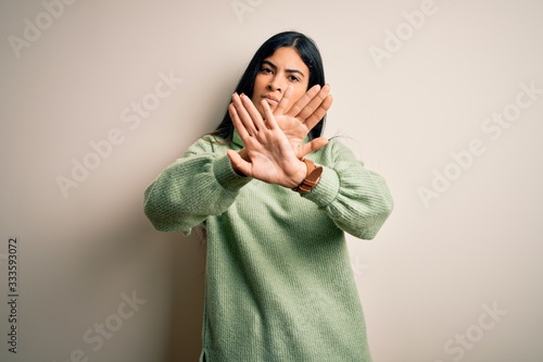 Young beautiful hispanic woman wearing green winter sweater over isolated background Rejection expression crossing arms and palms doing negative sign, angry face