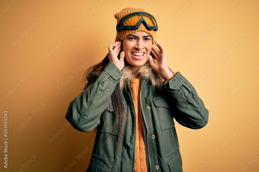 Young beautiful hispanic woman wearing ski glasses and coat for winter weather covering ears with fingers with annoyed expression for the noise of loud music. Deaf concept. Young beautiful hispanic woman wearing ski glasses and coat for winter weather covering ears with fingers with annoyed expression for the noise of loud music. Deaf concept.