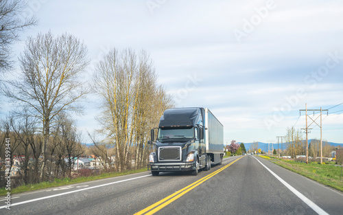 Black big rig semi truck transporting cargo in dry van semi trailer running on the two line road
