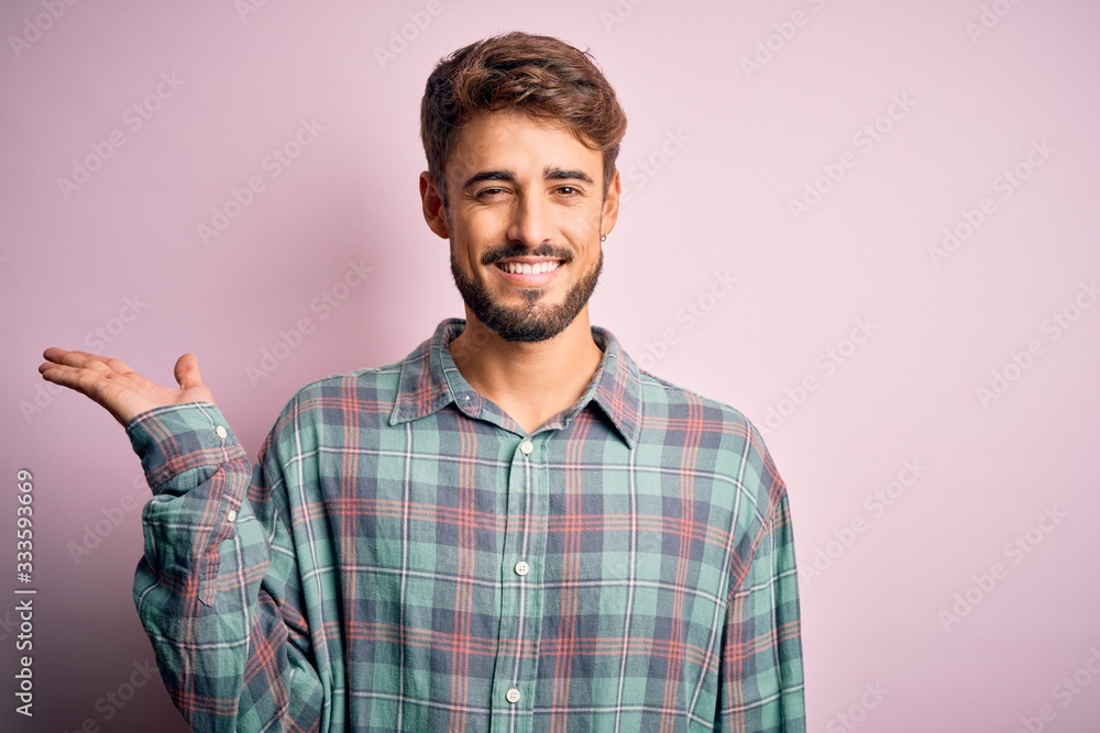 Young handsome man with beard wearing casual shirt standing over pink background smiling cheerful presenting and pointing with palm of hand looking at the camera. Young handsome man with beard wearing casual shirt standing over pink background smiling cheerful presenting and pointing with palm of hand looking at the camera.