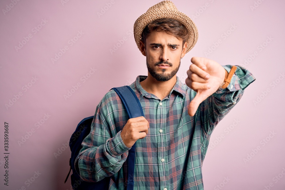 Young tourist man on vacation wearing backpack and summer hat over pink background with angry face, negative sign showing dislike with thumbs down, rejection concept