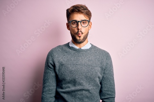Young handsome man with beard wearing glasses and sweater standing over pink background making fish face with lips, crazy and comical gesture. Funny expression. © Krakenimages.com