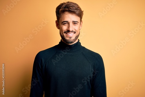 Canvas Print Young handsome man with beard wearing turtleneck sweater standing over yellow background with a happy and cool smile on face