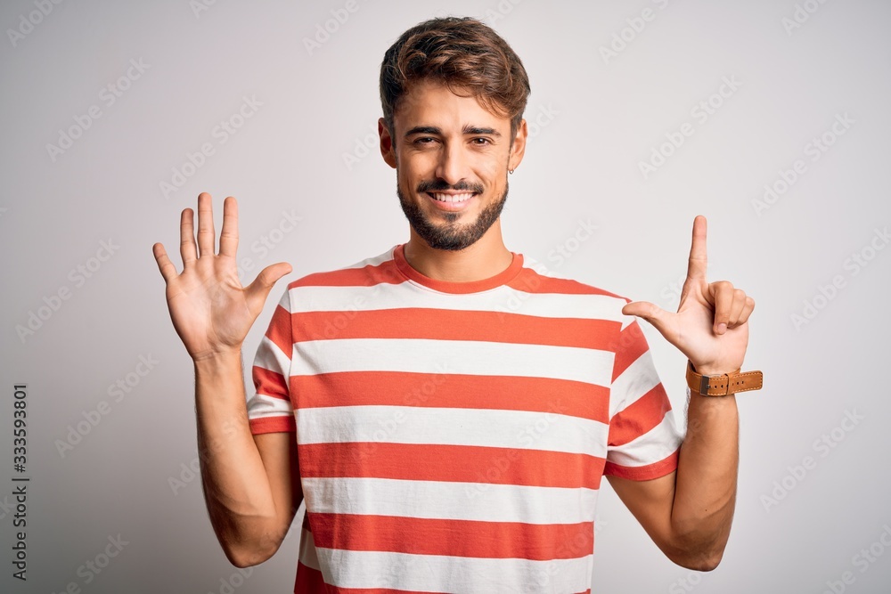 Young handsome man with beard wearing striped t-shirt standing over white background showing and pointing up with fingers number seven while smiling confident and happy. Young handsome man with beard wearing striped t-shirt standing over white background showing and pointing up with fingers number seven while smiling confident and happy.