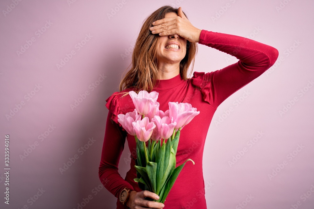 Young beautiful brunette woman holding bouquet of pink tulips over isolated background smiling and laughing with hand on face covering eyes for surprise. Blind concept.