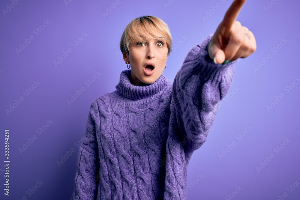 Young blonde woman with short hair wearing winter turtleneck sweater over purple background Pointing with finger surprised ahead, open mouth amazed expression, something on the front Young blonde woman with short hair wearing winter turtleneck sweater over purple background Pointing with finger surprised ahead, open mouth amazed expression, something on the front