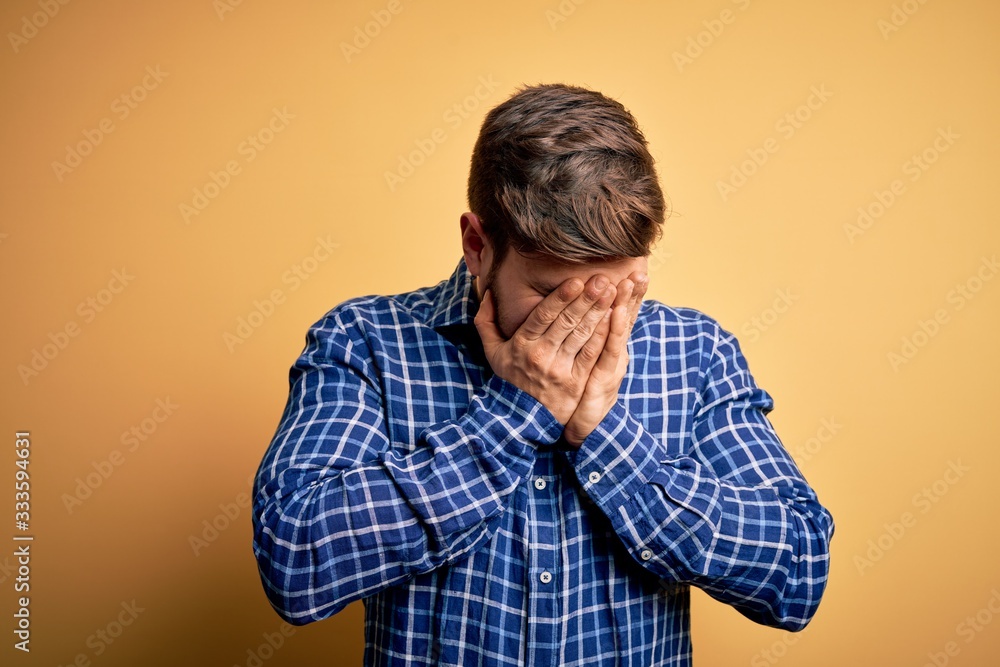 Young blond businessman with beard and blue eyes wearing shirt over yellow background with sad expression covering face with hands while crying. Depression concept. Young blond businessman with beard and blue eyes wearing shirt over yellow background with sad expression covering face with hands while crying. Depression concept.