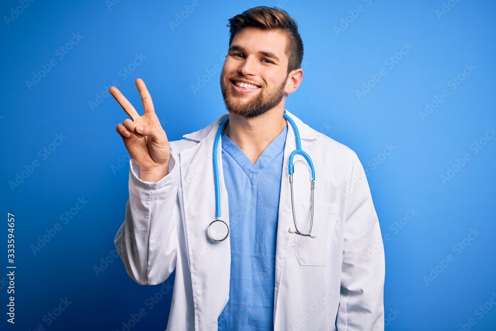 Young blond doctor man with beard and blue eyes wearing white coat and stethoscope showing and pointing up with fingers number two while smiling confident and happy. Young blond doctor man with beard and blue eyes wearing white coat and stethoscope showing and pointing up with fingers number two while smiling confident and happy.