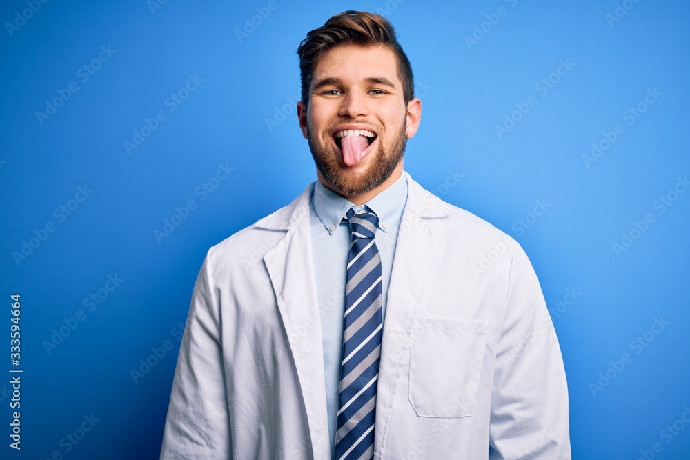 Young blond therapist man with beard and blue eyes wearing coat and tie over background sticking tongue out happy with funny expression. Emotion concept. Young blond therapist man with beard and blue eyes wearing coat and tie over background sticking tongue out happy with funny expression. Emotion concept.