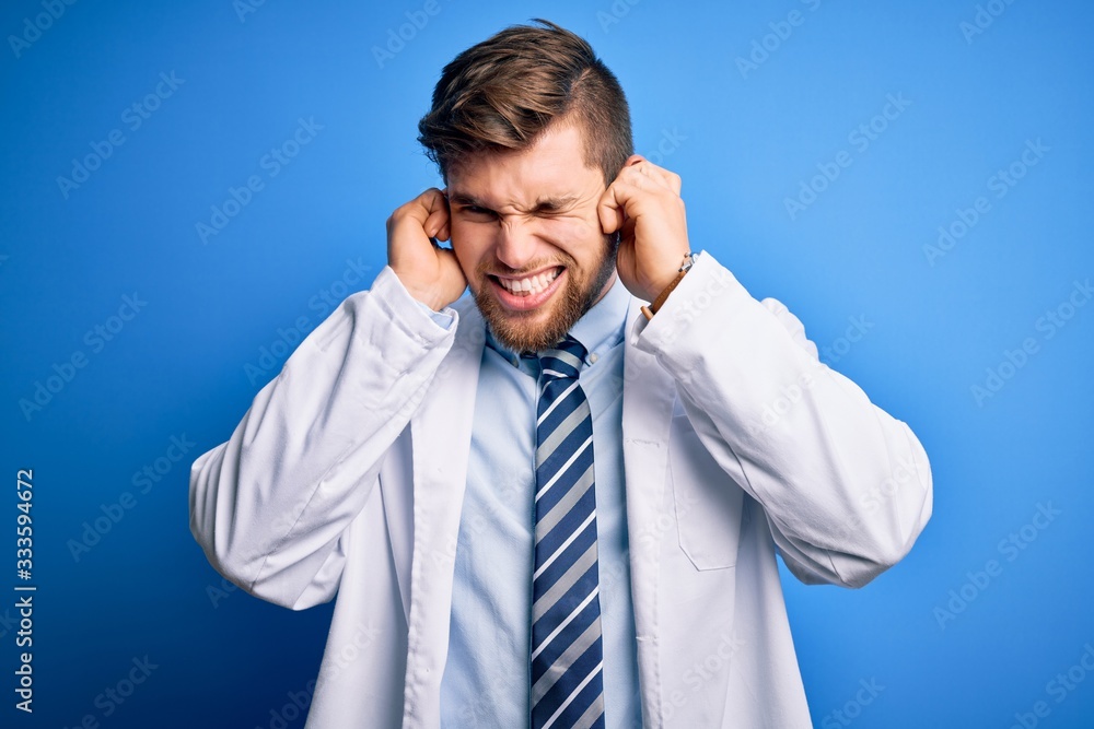 Young blond therapist man with beard and blue eyes wearing coat and tie over background covering ears with fingers with annoyed expression for the noise of loud music. Deaf concept. Young blond therapist man with beard and blue eyes wearing coat and tie over background covering ears with fingers with annoyed expression for the noise of loud music. Deaf concept.