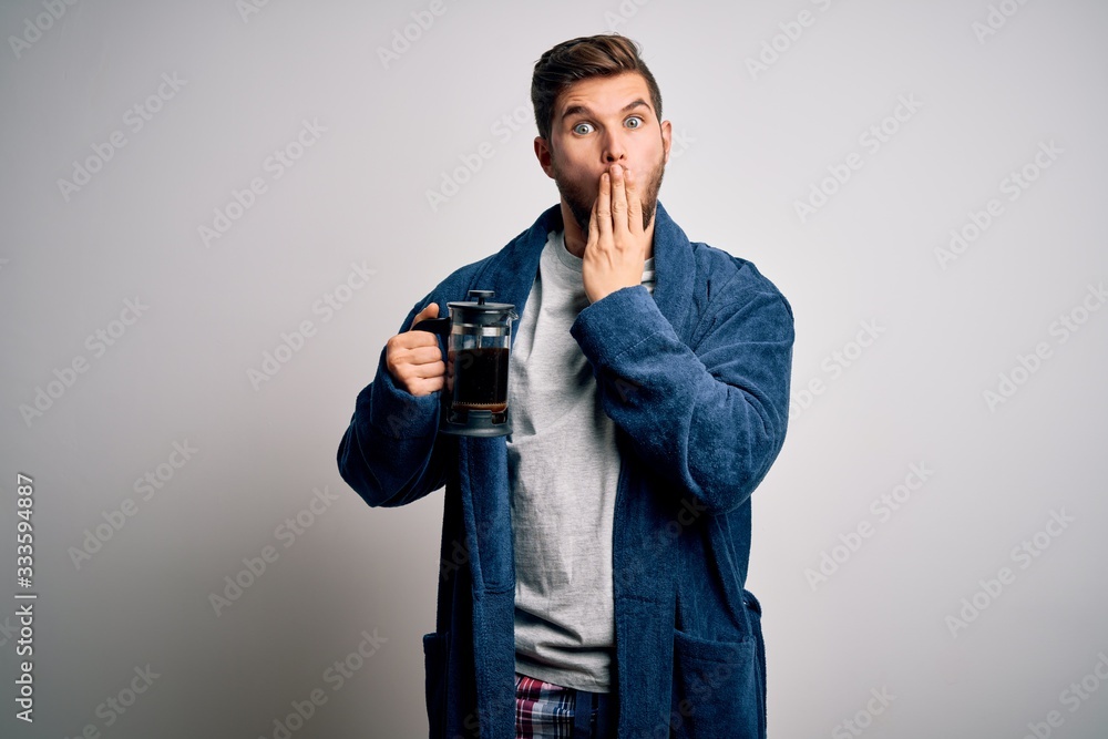 Young blond man with beard and blue eyes wearing pajama making coffe using coffeemaker cover mouth with hand shocked with shame for mistake, expression of fear, scared in silence, secret concept