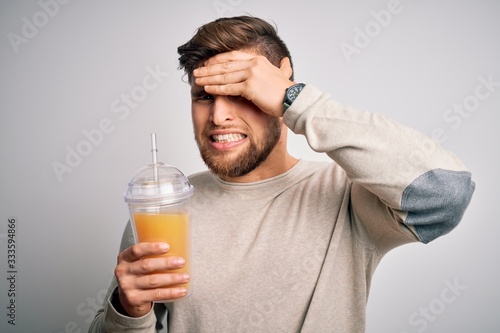 Young blond man with beard and blue eyes drinking healthy orange smoothie stressed with hand on head, shocked with shame and surprise face, angry and frustrated. Fear and upset for mistake.