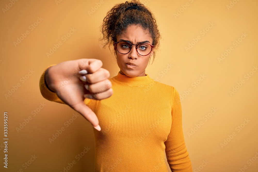 Young beautiful african american girl wearing sweater and glasses over yellow background looking unhappy and angry showing rejection and negative with thumbs down gesture. Bad expression. Young beautiful african american girl wearing sweater and glasses over yellow background looking unhappy and angry showing rejection and negative with thumbs down gesture. Bad expression.