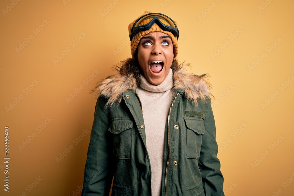 Young african american afro skier girl wearing snow sportswear and ski goggles angry and mad screaming frustrated and furious, shouting with anger. Rage and aggressive concept. Young african american afro skier girl wearing snow sportswear and ski goggles angry and mad screaming frustrated and furious, shouting with anger. Rage and aggressive concept.