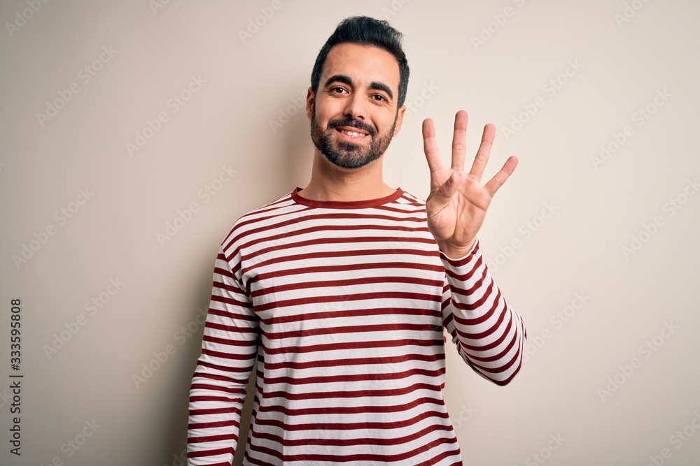 Naklejka premium Young handsome man with beard wearing casual striped t-shirt standing over white background showing and pointing up with fingers number four while smiling confident and happy.