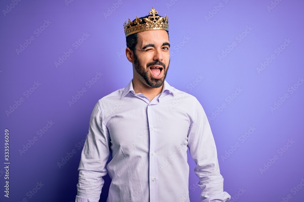 Young handsome man with beard wearing golden crown of king over purple background winking looking at the camera with sexy expression, cheerful and happy face.