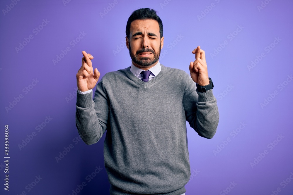 Handsome businessman with beard wearing casual tie standing over purple background gesturing finger crossed smiling with hope and eyes closed. Luck and superstitious concept. Handsome businessman with beard wearing casual tie standing over purple background gesturing finger crossed smiling with hope and eyes closed. Luck and superstitious concept.