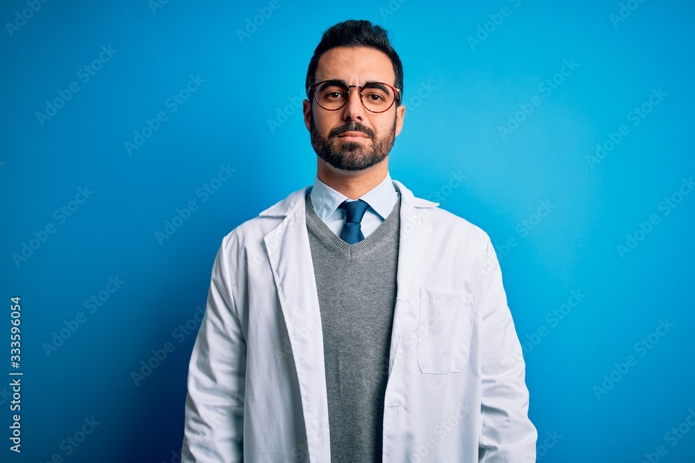 Young handsome doctor man with beard wearing coat and glasses over blue background Relaxed with serious expression on face. Simple and natural looking at the camera. Young handsome doctor man with beard wearing coat and glasses over blue background Relaxed with serious expression on face. Simple and natural looking at the camera.