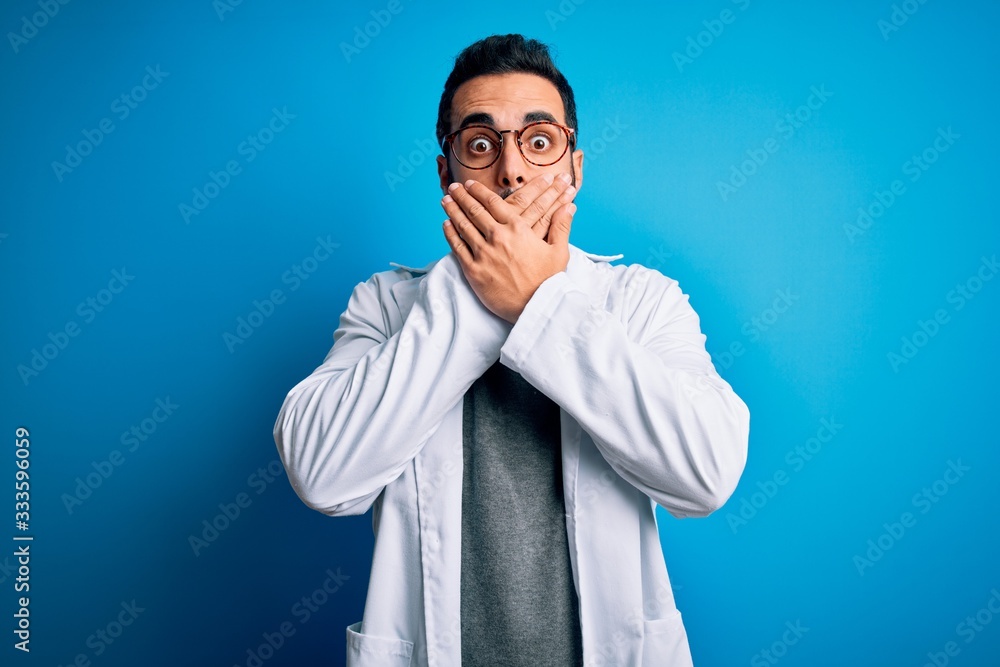 Young handsome doctor man with beard wearing coat and glasses over blue background shocked covering mouth with hands for mistake. Secret concept. Young handsome doctor man with beard wearing coat and glasses over blue background shocked covering mouth with hands for mistake. Secret concept.