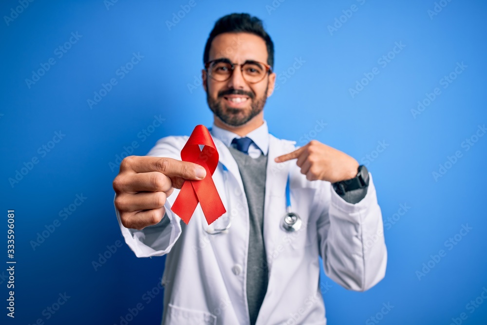 Young handsome doctor man with beard wearing stethoscope holding red hiv ribbon with surprise face pointing finger to himself Young handsome doctor man with beard wearing stethoscope holding red hiv ribbon with surprise face pointing finger to himself