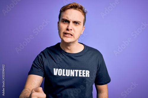 Young handsome redhead man wearing volunteer t-shirt over isolated purple background annoyed and frustrated shouting with anger, yelling crazy with anger and hand raised