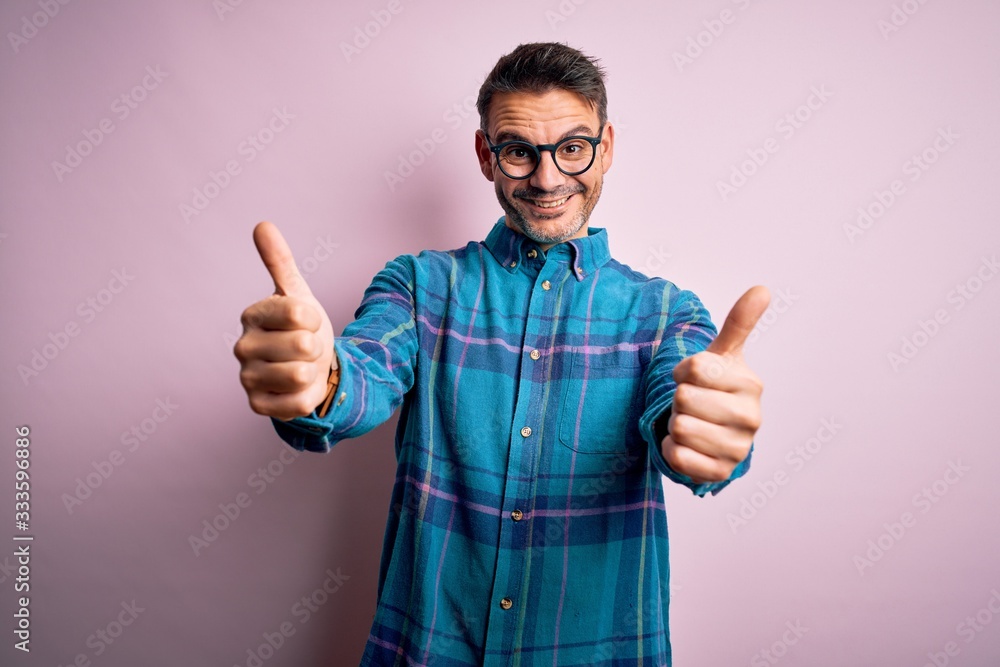 Young handsome man wearing casual shirt and glasses standing over isolated pink background approving doing positive gesture with hand, thumbs up smiling and happy for success. Winner gesture. Young handsome man wearing casual shirt and glasses standing over isolated pink background approving doing positive gesture with hand, thumbs up smiling and happy for success. Winner gesture.