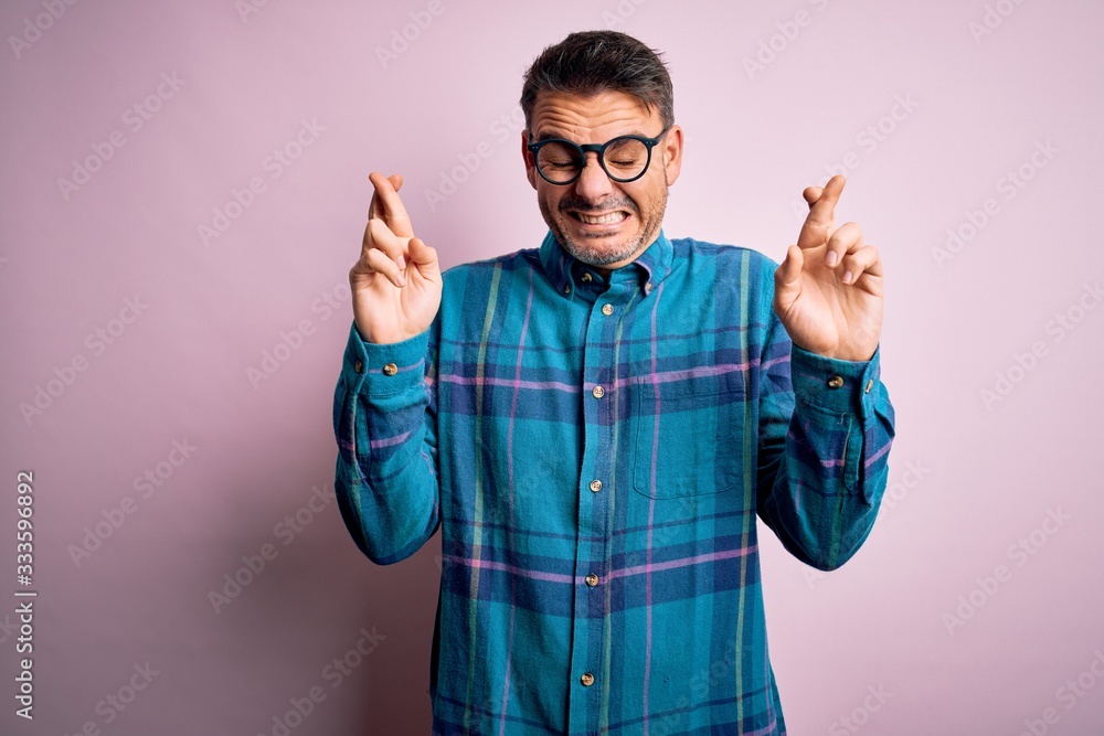 Young handsome man wearing casual shirt and glasses standing over isolated pink background gesturing finger crossed smiling with hope and eyes closed. Luck and superstitious concept. Young handsome man wearing casual shirt and glasses standing over isolated pink background gesturing finger crossed smiling with hope and eyes closed. Luck and superstitious concept.