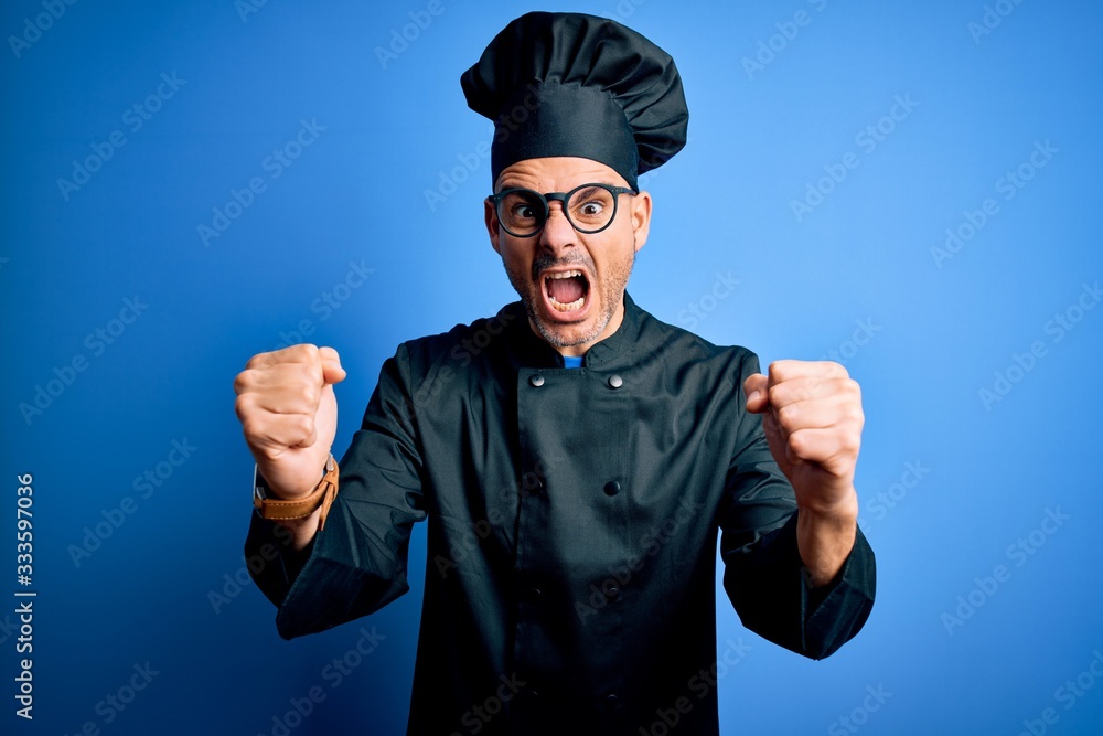 Young handsome chef man wearing cooker uniform and hat over isolated blue background angry and mad raising fists frustrated and furious while shouting with anger. Rage and aggressive concept. Young handsome chef man wearing cooker uniform and hat over isolated blue background angry and mad raising fists frustrated and furious while shouting with anger. Rage and aggressive concept.