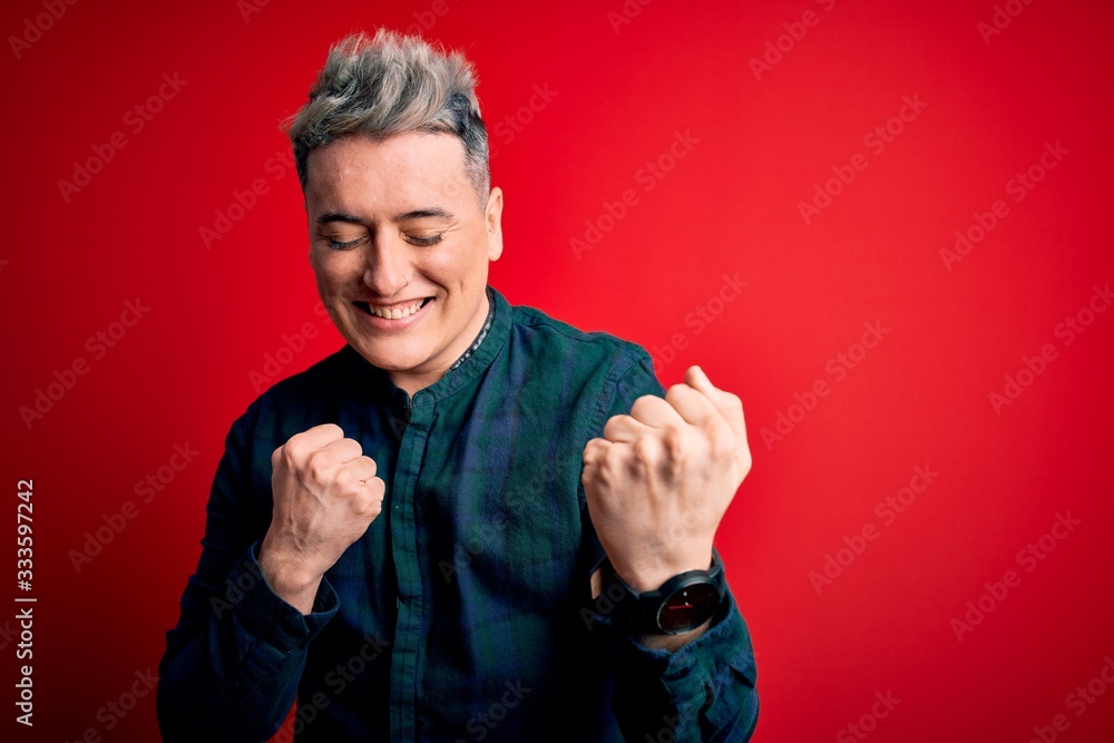 Young handsome modern man wearing elegant green shirt over red isolated background celebrating surprised and amazed for success with arms raised and eyes closed. Winner concept. Young handsome modern man wearing elegant green shirt over red isolated background celebrating surprised and amazed for success with arms raised and eyes closed. Winner concept.