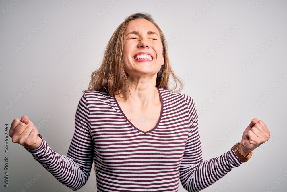 Young beautiful blonde woman wearing casual striped t-shirt over isolated white background very happy and excited doing winner gesture with arms raised, smiling and screaming for success. Celebration Young beautiful blonde woman wearing casual striped t-shirt over isolated white background very happy and excited doing winner gesture with arms raised, smiling and screaming for success. Celebration