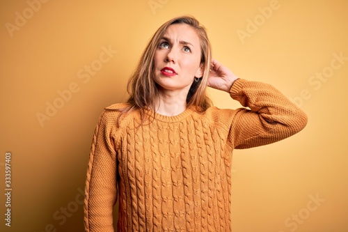 Young beautiful blonde woman wearing casual sweater standing over yellow background confuse and wondering about question. Uncertain with doubt, thinking with hand on head. Pensive concept. © Krakenimages.com