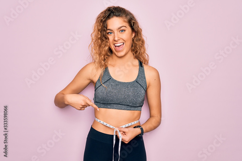 Young blonde sporty woman with blue eyes controlling weight using tape measure on waist very happy pointing with hand and finger © Krakenimages.com