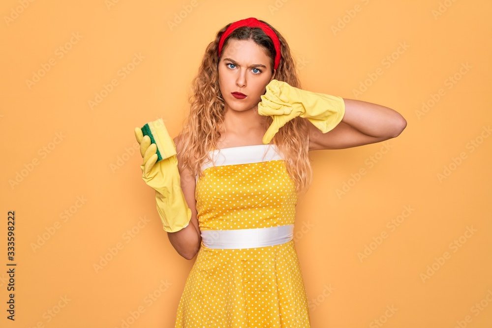 Young beautiful blonde woman with blue eyes cleaning using gloves and cleaner scourer with angry face, negative sign showing dislike with thumbs down, rejection concept Young beautiful blonde woman with blue eyes cleaning using gloves and cleaner scourer with angry face, negative sign showing dislike with thumbs down, rejection concept