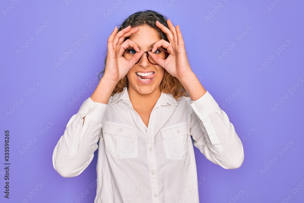 Young beautiful woman with blue eyes wearing casual shirt and glasses over purple background doing ok gesture like binoculars sticking tongue out, eyes looking through fingers. Crazy expression. Young beautiful woman with blue eyes wearing casual shirt and glasses over purple background doing ok gesture like binoculars sticking tongue out, eyes looking through fingers. Crazy expression.