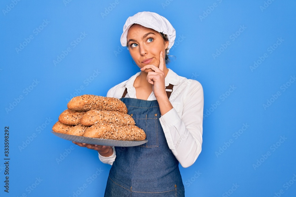 Young beautiful blonde baker woman with blue eyes holding tray with wholemeal bread cereal serious face thinking about question, very confused idea Young beautiful blonde baker woman with blue eyes holding tray with wholemeal bread cereal serious face thinking about question, very confused idea