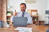 Middle age handsome businessman wearing tie sitting using laptop at the office doing happy thumbs up gesture with hand. Approving expression looking at the camera with showing success.
