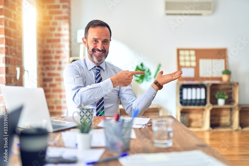 Middle age handsome businessman wearing tie sitting using laptop at the office amazed and smiling to the camera while presenting with hand and pointing with finger.