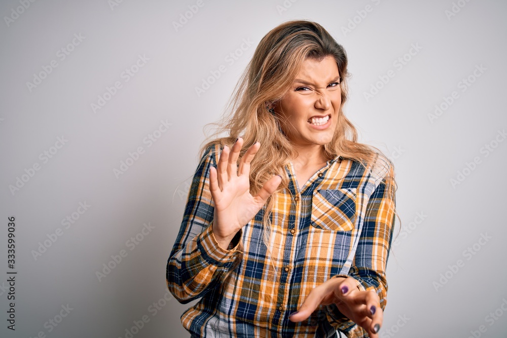 Young beautiful blonde woman wearing casual shirt standing over isolated white background disgusted expression, displeased and fearful doing disgust face because aversion reaction. With hands raised Young beautiful blonde woman wearing casual shirt standing over isolated white background disgusted expression, displeased and fearful doing disgust face because aversion reaction. With hands raised