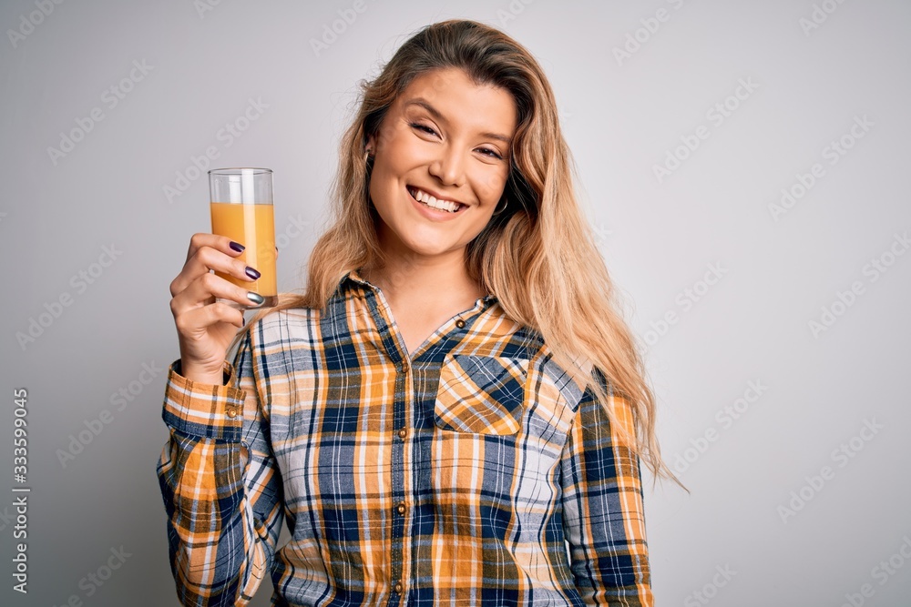Young beautiful blonde woman drinking glass of healthy orange juice over white background with a happy face standing and smiling with a confident smile showing teeth Young beautiful blonde woman drinking glass of healthy orange juice over white background with a happy face standing and smiling with a confident smile showing teeth