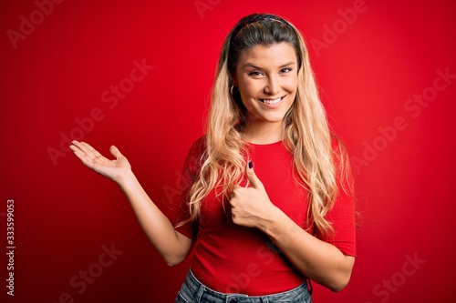 Young beautiful blonde woman wearing casual t-shirt standing over isolated red background Showing palm hand and doing ok gesture with thumbs up, smiling happy and cheerful