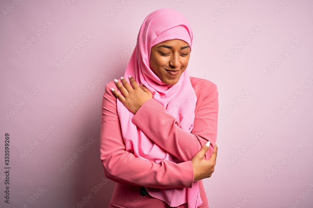 Young African American afro woman wearing muslim hijab over isolated pink background Hugging oneself happy and positive, smiling confident. Self love and self care Young African American afro woman wearing muslim hijab over isolated pink background Hugging oneself happy and positive, smiling confident. Self love and self care