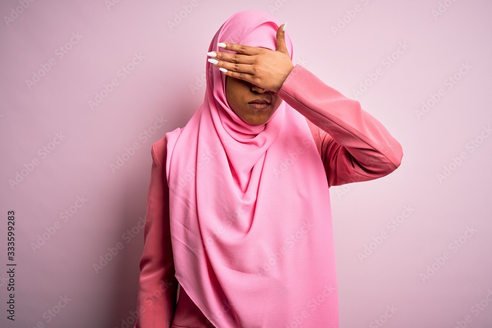 Young African American afro woman wearing muslim hijab over isolated pink background covering eyes with hand, looking serious and sad. Sightless, hiding and rejection concept Young African American afro woman wearing muslim hijab over isolated pink background covering eyes with hand, looking serious and sad. Sightless, hiding and rejection concept