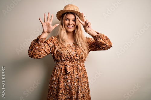 Young beautiful blonde woman wearing summer dress and hat over isolated white background showing and pointing up with fingers number seven while smiling confident and happy.