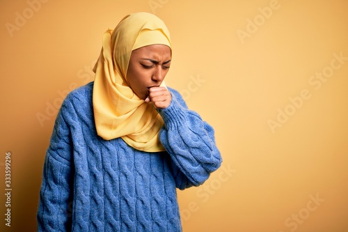 Young African American afro woman wearing muslim hijab over isolated yellow background feeling unwell and coughing as symptom for cold or bronchitis. Health care concept.