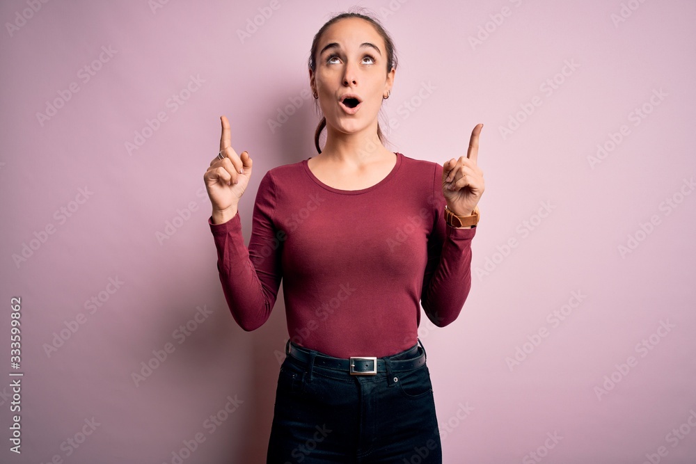Young beautiful woman wearing casual t-shirt standing over isolated pink background amazed and surprised looking up and pointing with fingers and raised arms. Young beautiful woman wearing casual t-shirt standing over isolated pink background amazed and surprised looking up and pointing with fingers and raised arms.